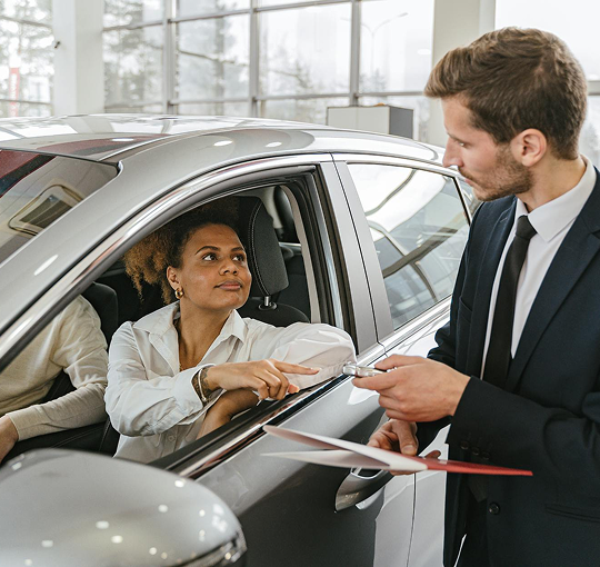Eine Frau sitzt in einem Auto, während ein Verkäufer ihr Informationen gibt und ein Dokument hält.