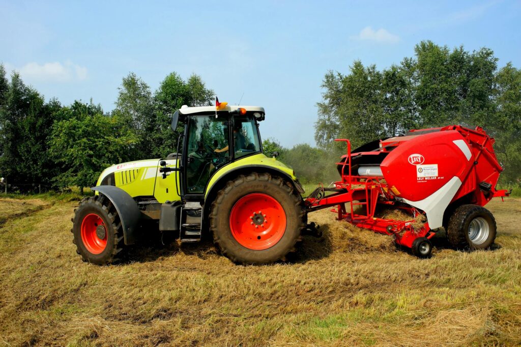 Ein grüner Traktor mit rotem Anhänger bearbeitet ein Feld unter blauem Himmel und umgeben von Bäumen.