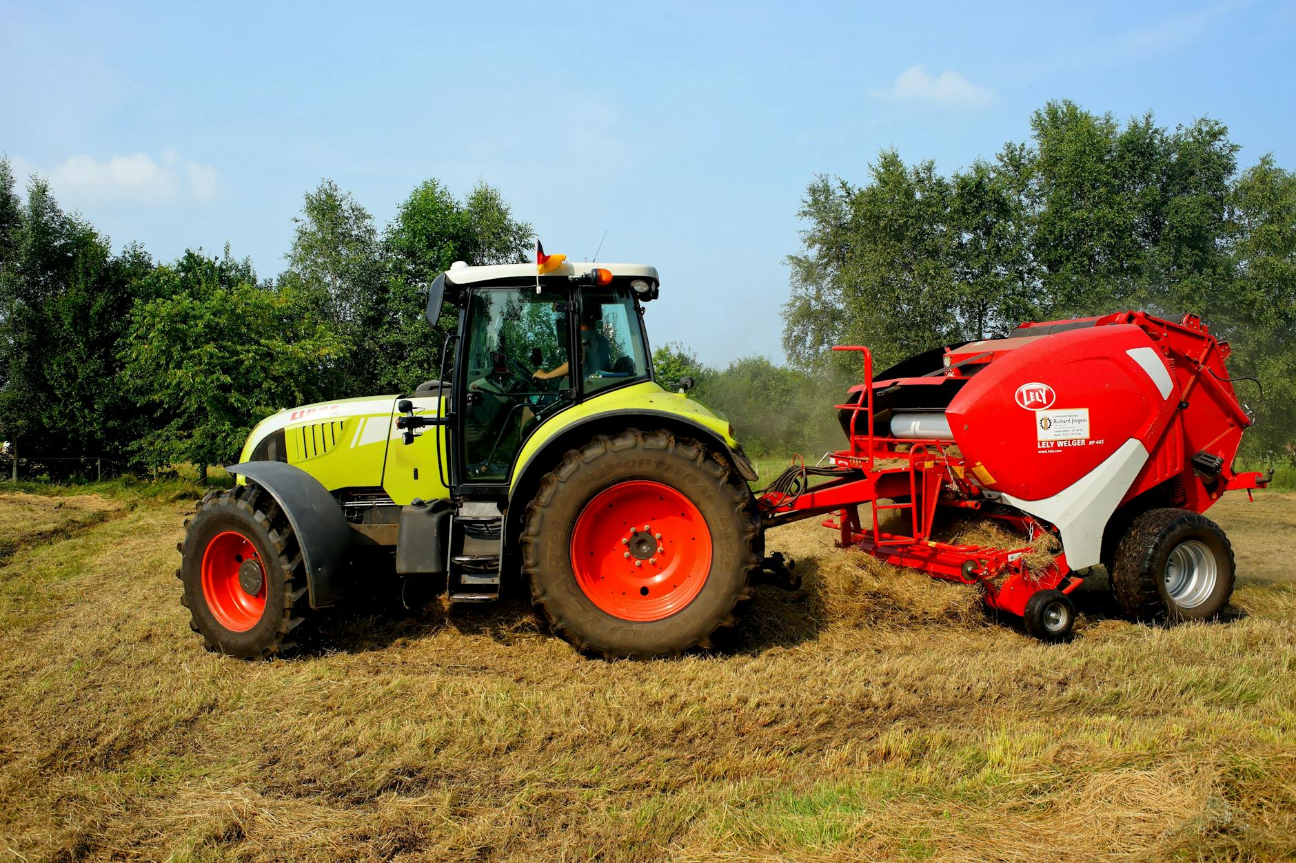 Ein grüner Traktor mit rotem Anhänger bearbeitet ein Feld unter blauem Himmel und umgeben von Bäumen.