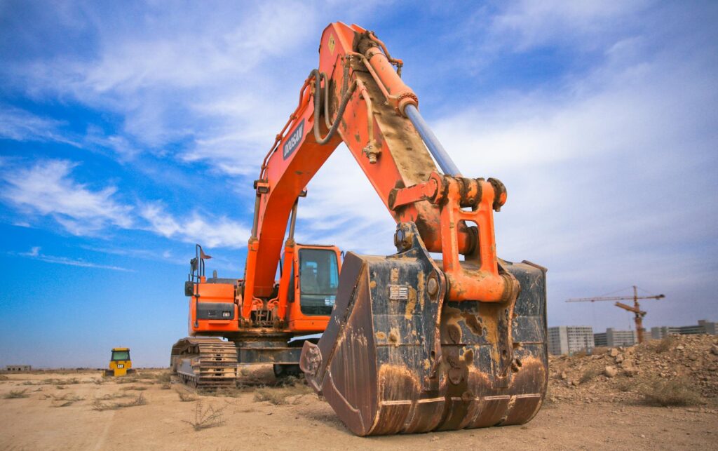 Ein orangefarbener Bagger steht im Vordergrund auf einer unebenen, sandigen Baustelle unter einem blauen Himmel.