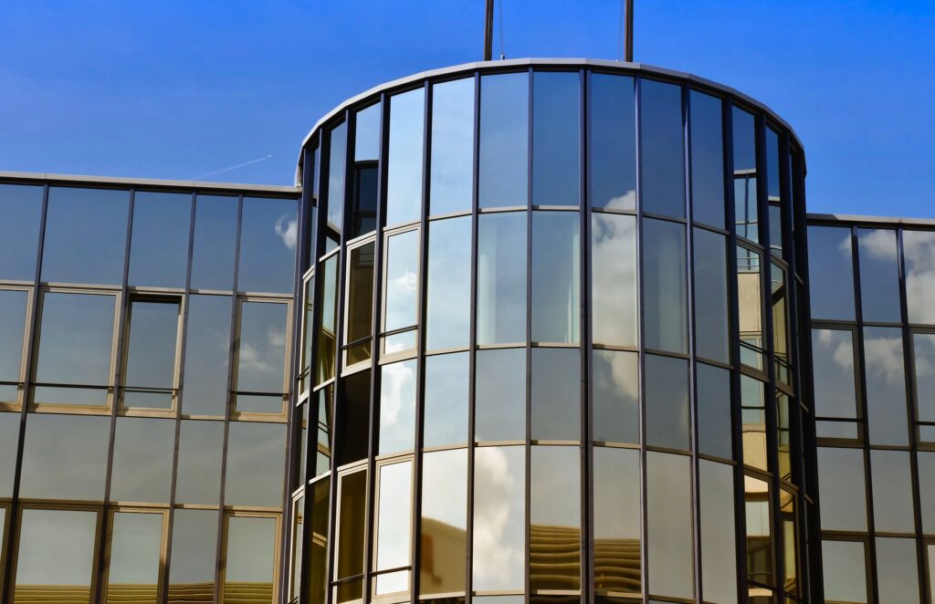 Moderne Glasfassade eines Gebäudes mit reflektierenden Fenstern und einem blauen Himmel im Hintergrund.
